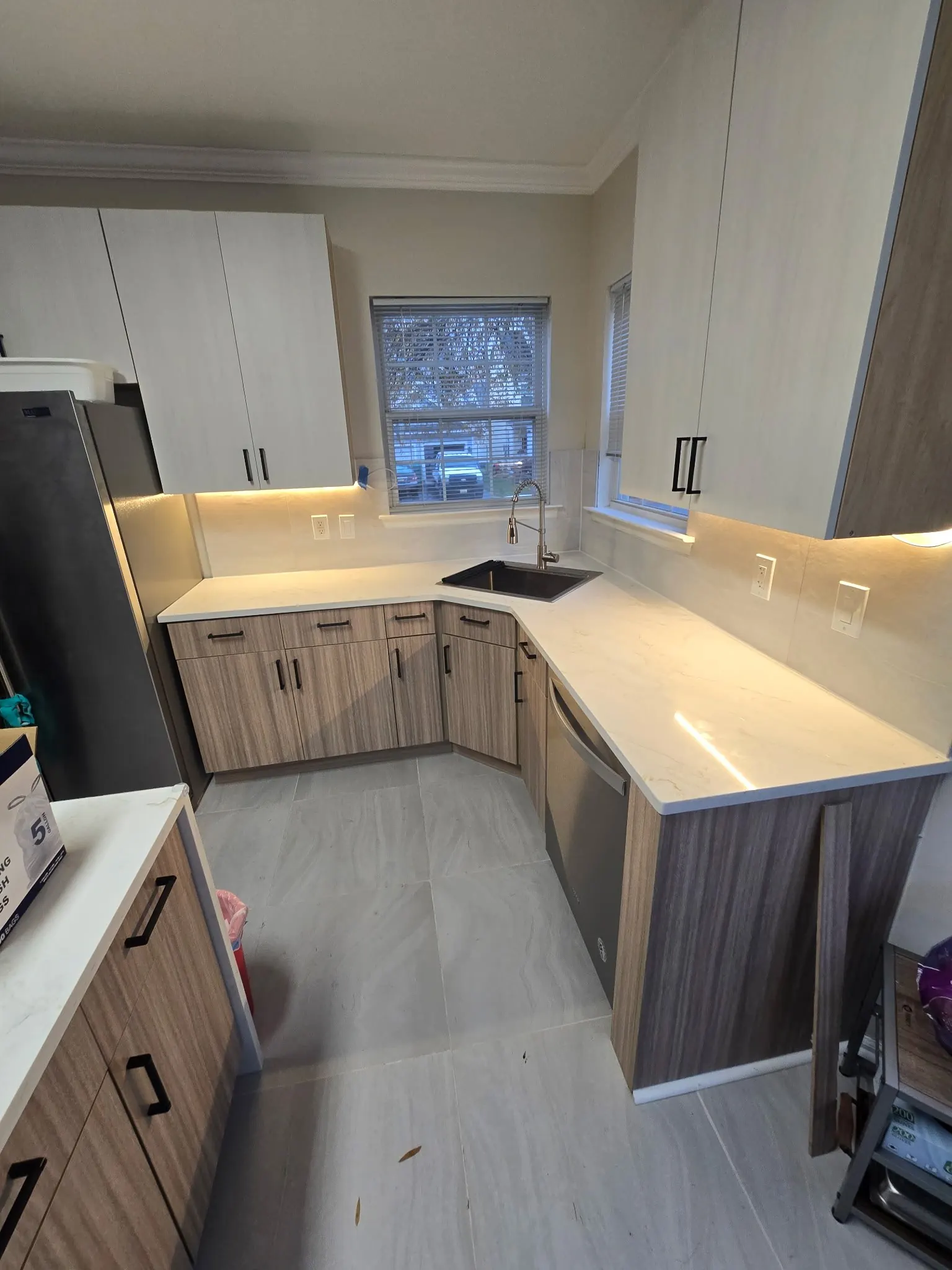 Completed kitchen with light upper cabinets, wood-grain lower cabinets, pale counters and under-cabinet lighting beside a corner window.