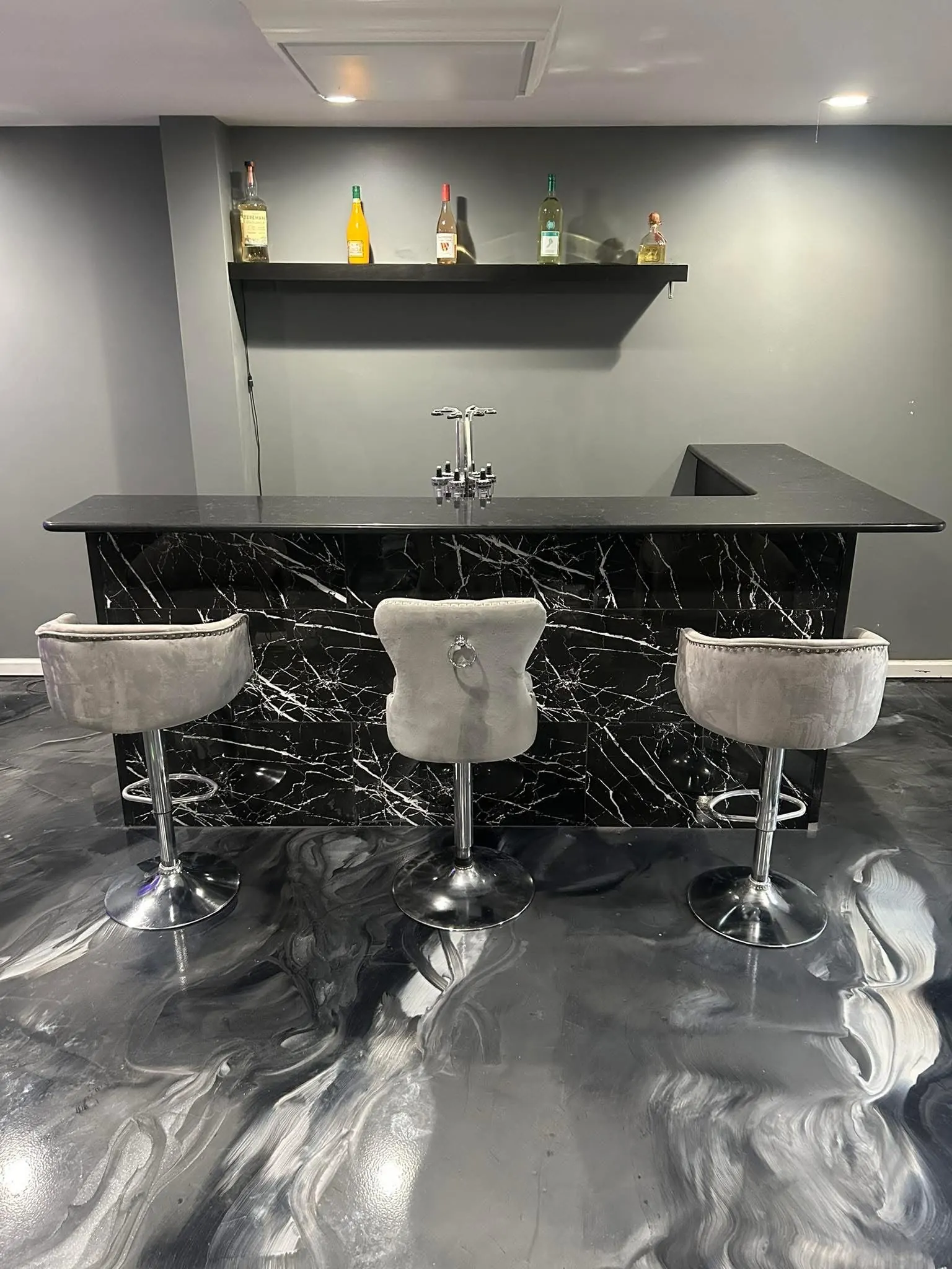 A bar with a dark veined surface, three gray stools and patterned flooring.